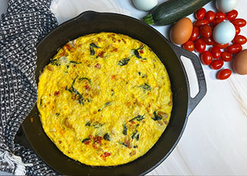 A golden spinach and white bean frittata with turmeric and tomato is served in a cast iron skillet with fresh eggs and veggies on the side.