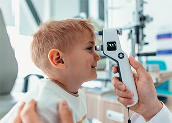 A five-year-old, smiling blond boy is getting his eyes checked with an ophthalmoscope.