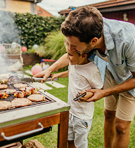 A dad is teaching his son how to turn food on the barbecue.
