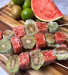 A trio of frozen watermelon kiwi skewers sprinkled with chili-lime salt are displayed on a wood cutting board with a fresh wedge of watermelon in the background.