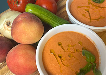 Two white bowls filled with tomato, peach and basic gazpacho are surrounded by fresh tomatoes, peaches and basil on a wood cutting board.