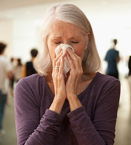 Silver-haired woman wearing a purple sweater sneezes into a tissue in a large room with several people in the background.