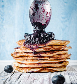 A stack of lemon ricotta pancakes sits on a gray board as a spoon tops them with a big dollop of blueberry compote.