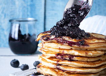 A  jam jar containing blueberry compote sits in the background while a big stack of lemon ricotta pancakes is being served.
