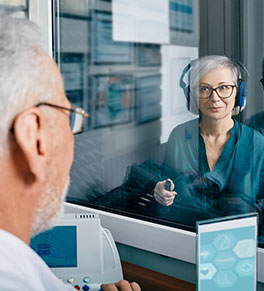 An attractive middle-aged woman in a teal blouse is taking an audiology test with older doctor seen through an observation window.