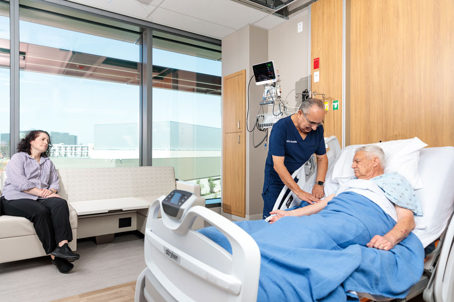 a nurse checks on the pulse of a patient while the patient's daughter sits in on a comfortable chair in one of the patient rooms at uci health — irvine