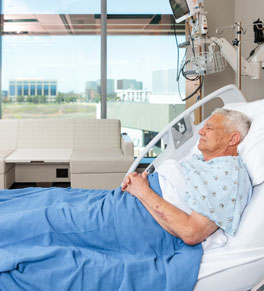 a patient at uci health irvine rests under a blue blanket near floor to ceiling windows
