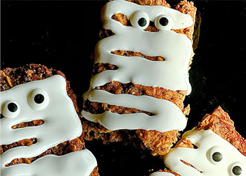 Pumpkin-spiced mummy bars are displayed on a black background with striped icing and googly eyes.