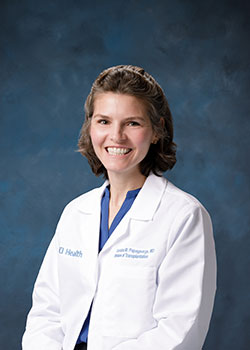 UCI Health transplant surgeon Dr. Christina Papageorge is sitting in her white coat against a dark blue background.
