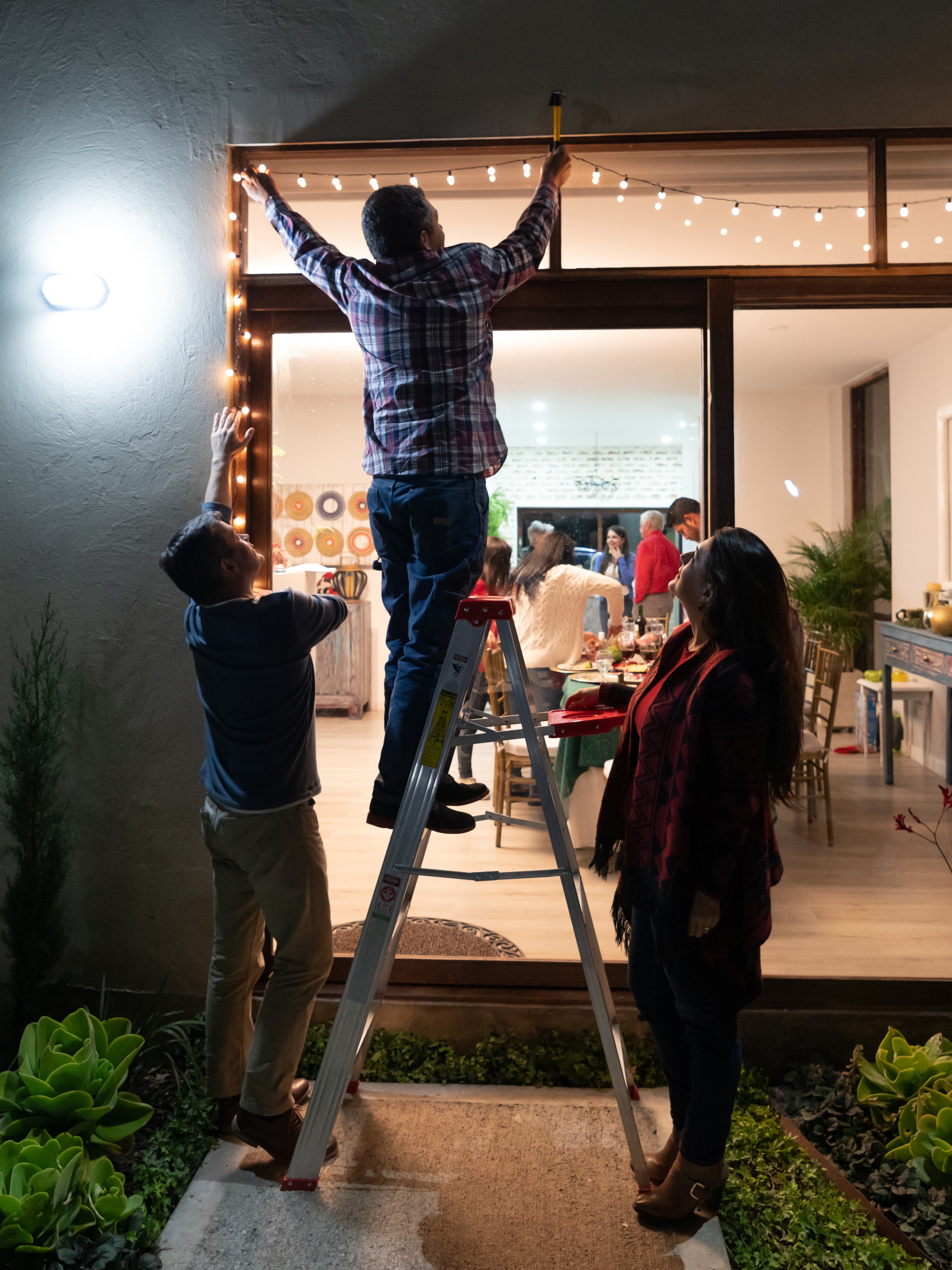 a man strings lights while standing on a ladder and being watched by two friends standing on the ground