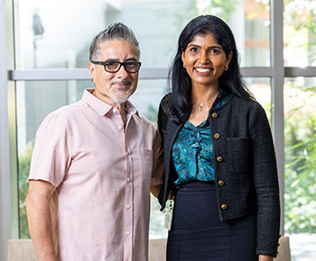 Gastric cancer survivor Mauricio Alvarez, pictured in a pink sport shirt at left, poses with his UCI Health surgical oncologist Dr. Maheswari Senthil, who is wearing a black skirt and sweater over a  turquoise print blouse.