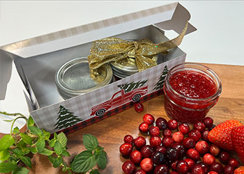 A plaid gift box with two jam jars sits next to an open jar of Christmas jam with fresh mint and cranberries in the foreground.