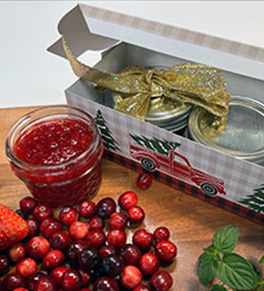 An open jar of Christmas jam sits near a gift box with two other jars surrounded by fresh cranberries and mint.