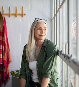 A smiling, mature woman with long gray hair and glasses atop her head looks out a window.