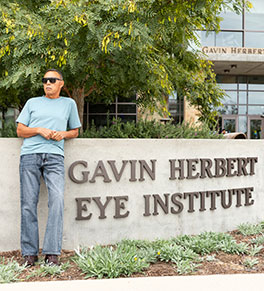 Sergio Reyes, wearing sunglasses, is leaning against the Gavin Herbert Eye Institute sign.