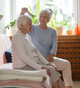 A older woman with dementia is sitting on a sofa while a smiling caregiver brushes her hair.