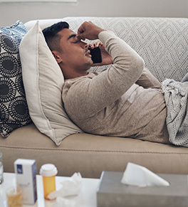 A sick young man is laying on a sofa talking on his cell phone and holding his forehead.