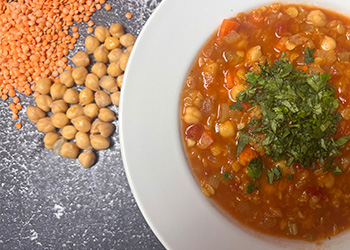 Creamy red Moroccan chickpea harira is served in a white bowl on a gray concrete counter with fresh chickpeas and red lentils spread to the left.