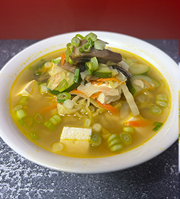 Hearty vegetable and chicken miso soup is stacked high in a white bowl in front of a red background.