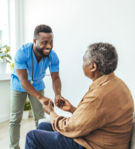 a male caregiver wearing scrubs and stethoscope smiles and helps a seated elderly patient stand up 