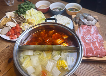 A split broth pot with two broths is sitting on a table surrounding by hot pot ingredients including vegetables, meat, seafood and sauces.