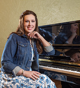 A smiling Kimberly Renihan sits by her Yamaha upright piano wearing a blue and white floral dress and blue jean jacket.