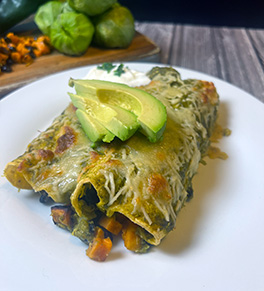 Black bean and sweet potato enchiladas are served on a white plate with avocado slices on top and tomatillos in the background.