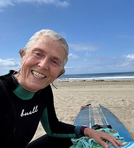 Spine patient Diane O'Dowd is fresh out of the water after surfing is smiling next to her blue surfboard.
