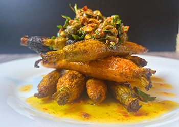 Rainbow carrots with harissa, herbs and pistachios are stacked crossways in a tower on a white plate with yellow sauce pooling.