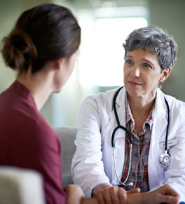 a female doctor in a white coat and stethoscope speaks to a woman while holding her hand