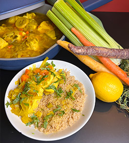 Poached chicken in a saffron, leek and parsley broth is served over farro on a white plate with a blue pot full of the entree in the background.