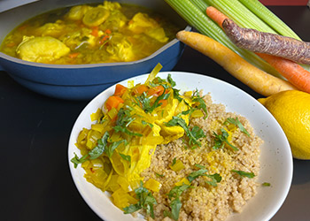 Poached chicken in saffron, leek and parsley broth is served over farro in a white bowl, a blue pot of the entree is in the background with whole vegetables on the side.