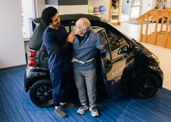 A rehabilitation staff member helps an older man out of a Smart car simulator.