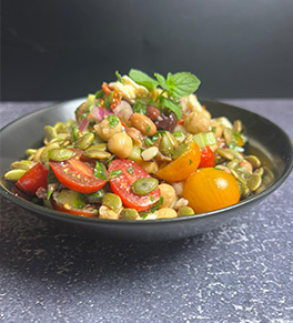 A black bowl is full of Mediterranean bean salad that is garnished with red and orange tomatoes and a sprig of mint.