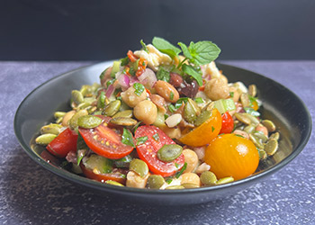 A heaping helping of Mediterranean bean salad is served in a black bowl that is resting on a blue textured countertop.
