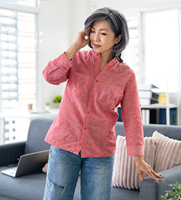 An middle-aged Asian woman is holding her head and leaning one hand on a blue sofa for support because of headache and dizziness.