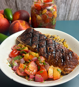 Cocoa-chili rubbed salmon is display with peach salsa in a white pasta bowl. Fresh peaches and a jar of salsa are in the background.