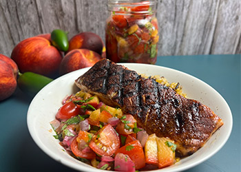 Grilled cocoa-chili rubbed salmon is served in a white pasta bowl alongside peach salsa. It sits on a turquoise counter with fresh peaches and a jar of salsa in the background.