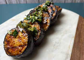 Five miso-maple glazed eggplant steaks drizzled with scallion gremolata are lined up on a white marble cutting board.