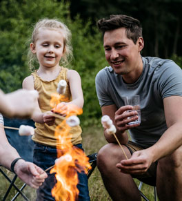 a father and daughter roasting marshmallows around a campfire with several other people's hands in the frame
