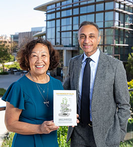 Janice Munemitsu, left, stands with her eye surgeon, Dr. Sanjay Kedhar, outside the UCI Health Gavin Herbert Eye Institute, The author is holding a copy of her book about school desegregation in Orange County.