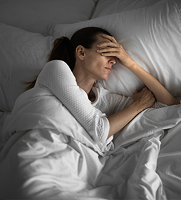 In a dimly lit bedroom, a woman lays in an all white bed, holding her head and covering her eyes because of migraine pain.