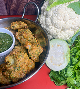 Air-fried cauliflower pakoras are displayed in a stainless steel wok with guava mint chutney on the side. The countertop is red and a head of fresh cauliflower, half a guava and cilantro are also displayed.
