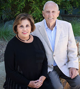 Renee Horner, at left, wearing a black top, sits with her husband Peter Horner, wearing a tan sport coat, sit together in a garden setting.