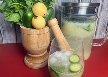 A sparkling guava cucumber mocktail is served in a wine glass, with a full pitcher, fresh veggies and wood pestle in the background.