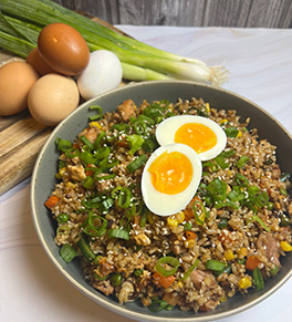 Any grain chicken fried rice is served in a gray bowl with scallions and fresh eggs on a bamboo cutting board in the background.