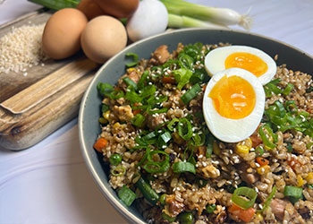 Any grain chicken fried rice is served in a gray bowl displayed with raw grains, fresh eggs and scallions on a cutting board to the left side.