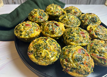 A dozen grain and egg muffins with spinach, feta and sun-dried tomato are displayed on a black plate with a dark green napkin tucked behind.