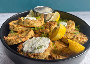 Lemon pepper salmon cakes are displayed on a black platter with yogurt-dill sauce and lemon wedge garnishes.