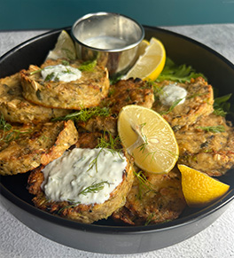 A black plate holds lemon pepper salmon cakes served with yogurt-dill sauce and fresh lemon slices for garnish.